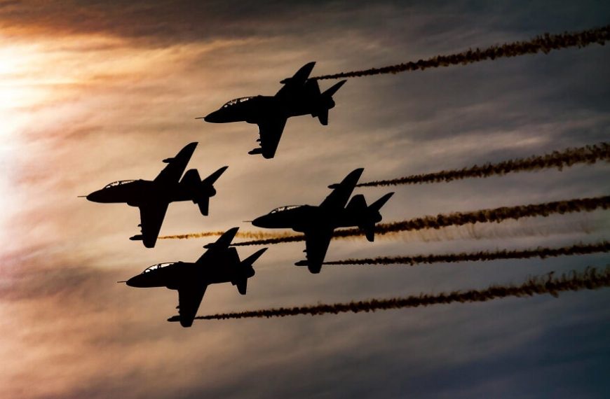 Four jet aircraft silhouetted in formation against a low sun and high cloud